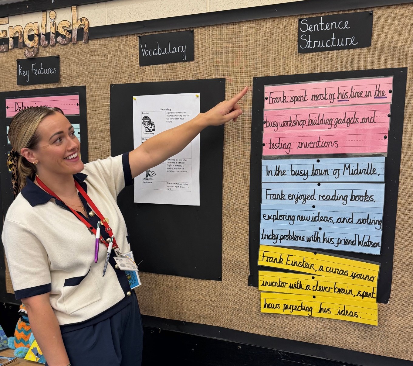 A primary school teacher smiles and points to a colourful classroom display on sentence structure and vocabulary, featuring pupil writing.