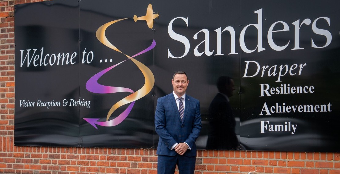 Stuart standing outside Sanders Draper School in front of the school's sign.