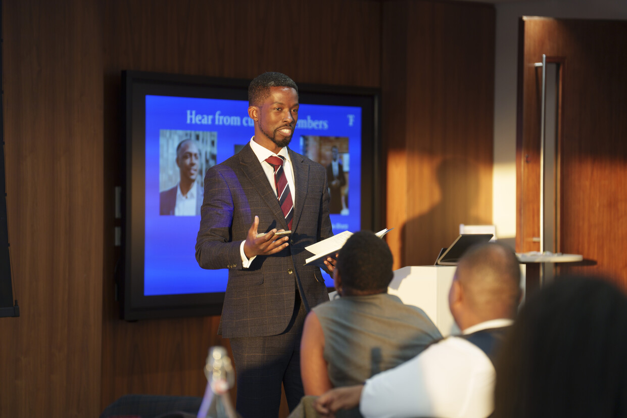 Michael, member of the 100 Black Heads Network, is standing and confidently delivering a presentation to peers at a national conference. He is dressed smartly in a grey suit, white shirt, and red and black striped tie. The three audience members in the foreground are watching attentively.