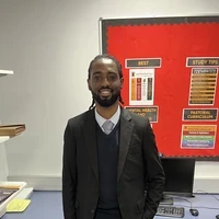 Jeffrey, a SCITT trainee teacher at Eastbrook School, standing indoors in a classroom setting, wearing professional attire, with a classroom display board visible behind him.