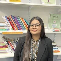 A maths teacher from a SCITT programme stands in a school resource area in front of shelves filled with educational books and teaching materials.