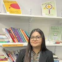 A maths teacher from a SCITT programme stands in a school resource area in front of shelves filled with educational books and teaching materials.