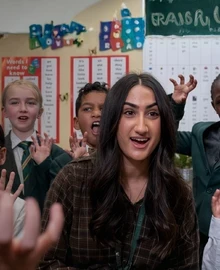 A teacher leads an interactive classroom activity with a group of engaged primary school pupils, hands raised.