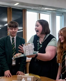 A teacher shows a small architectural model to a group of secondary school pupils in green uniform, who react with delight.
