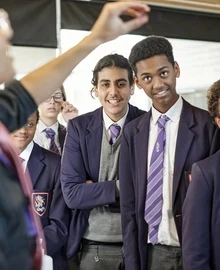 A group of secondary school pupils in uniform stand together in a classroom, smiling."