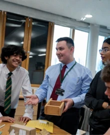 A teacher shows a wooden box to a group of engaged secondary school pupils around a table in a design and technology classroom.