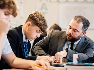 Teacher in a suit helping a male student with a maths problem in a classroom setting