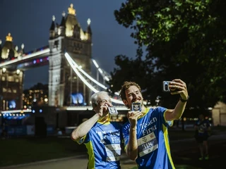 Two smiling runners wearing Teach First shirts take a selfie with their medals in front of Tower Bridge at night.