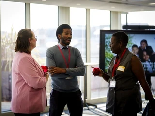 People talking at a Teach First event in a bright office space. Three colleagues wearing lanyards stand in conversation, holding drinks, with a presentation screen in the background.