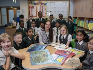 A teacher sits with a group of primary school pupils around a table, looking at books together in a classroom.