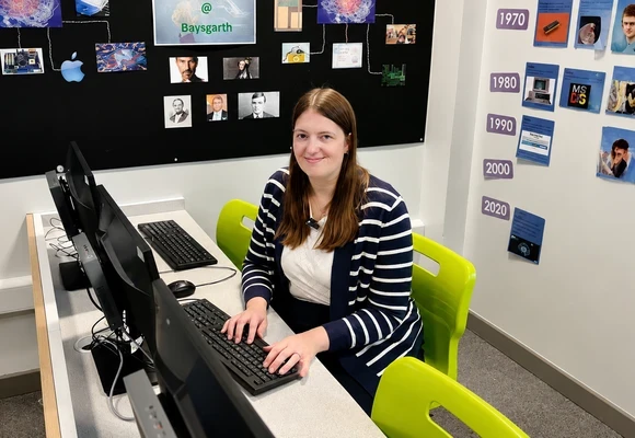 Sarah using a computer in a classroom with technology displays and timelines on the walls.