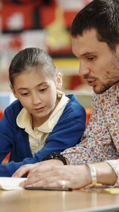 A teacher seated at a classroom table reading a book with two pupils.