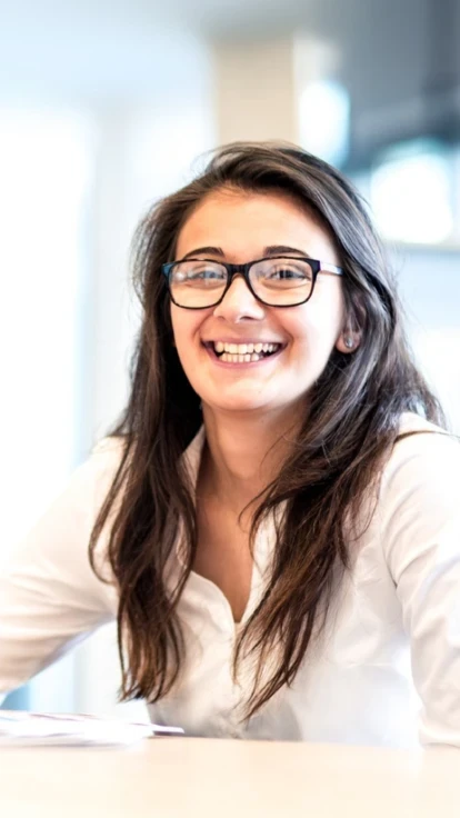 Female teacher smiling enthusiastically while writing with a pen at a desk. She wears rectangular glasses and has long straight brownish-black hair.