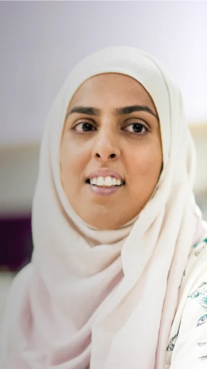 Smiling woman in a headscarf sitting in a professional setting, with a blurred background. Related to Headship Next programme.