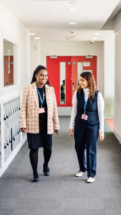 Two staff members walking side by side down a bright school corridor, smiling and talking, with lockers along the wall and a red door in the background.