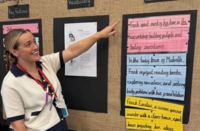 A primary school teacher smiles and points to a colourful classroom display on sentence structure and vocabulary, featuring pupil writing.