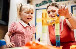 A primary school-aged girl and an adult teacher explore a marble run toy together in a classroom setting, focusing on teamwork and learning through play.
