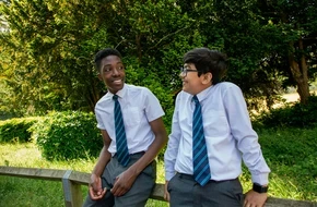Two secondary school boys in uniform sitting outside, smiling and talking in a green, sunny setting.