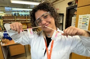 Fatima, wearing a lab coat and safety goggles, holds up results from a science experiment in a classroom laboratory.