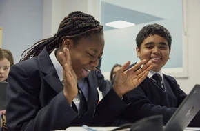 Two secondary school students in uniform smiling and laughing together while using a laptop in a classroom.