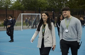 Two teachers wearing lanyards walking across a school playground with pupils in the background.