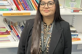 A maths teacher from a SCITT programme stands in a school resource area in front of shelves filled with educational books and teaching materials.