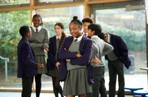 Secondary school pupils in uniform socialising indoors, with one pupil standing confidently in the foreground.