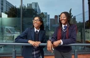 Two secondary school girls in uniform standing outside their school, smiling and looking ahead