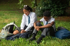Two secondary school pupils in uniform sitting together on grass outside school.