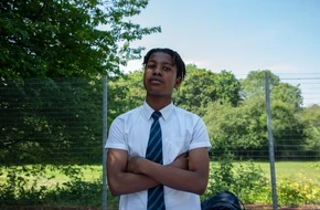 Secondary school pupil in uniform standing outdoors with arms folded in front of a fenced sports area