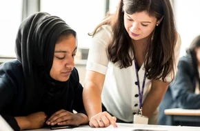 A teacher supports a pupil with her work at a desk in a classroom.