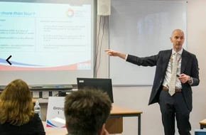 Tim, a teacher, presenting to students in a classroom with a slide projected behind him.