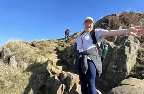 Fran smiling with her arms outstretched while walking on a rocky hillside under a bright blue sky.