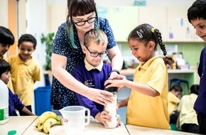 A teacher helps primary school pupils prepare a drink using a blender during a classroom activity.