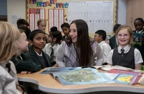 A smiling teacher sits with a group of primary school pupils around a table of open picture books in a colourful classroom.