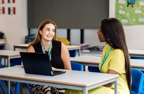 Two women sitting at a classroom table, smiling and talking, with an open laptop in front of them.