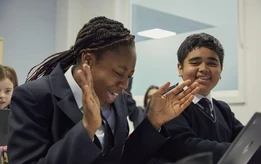 Two secondary school students in uniform smiling and laughing together while using a laptop in a classroom.