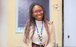Smiling teacher wearing glasses and a lanyard sitting in a classroom doorway.