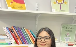 A maths teacher from a SCITT programme stands in a school resource area in front of shelves filled with educational books and teaching materials.