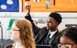 Secondary school pupil raising his hand during a classroom lesson, with other pupils seated nearby.